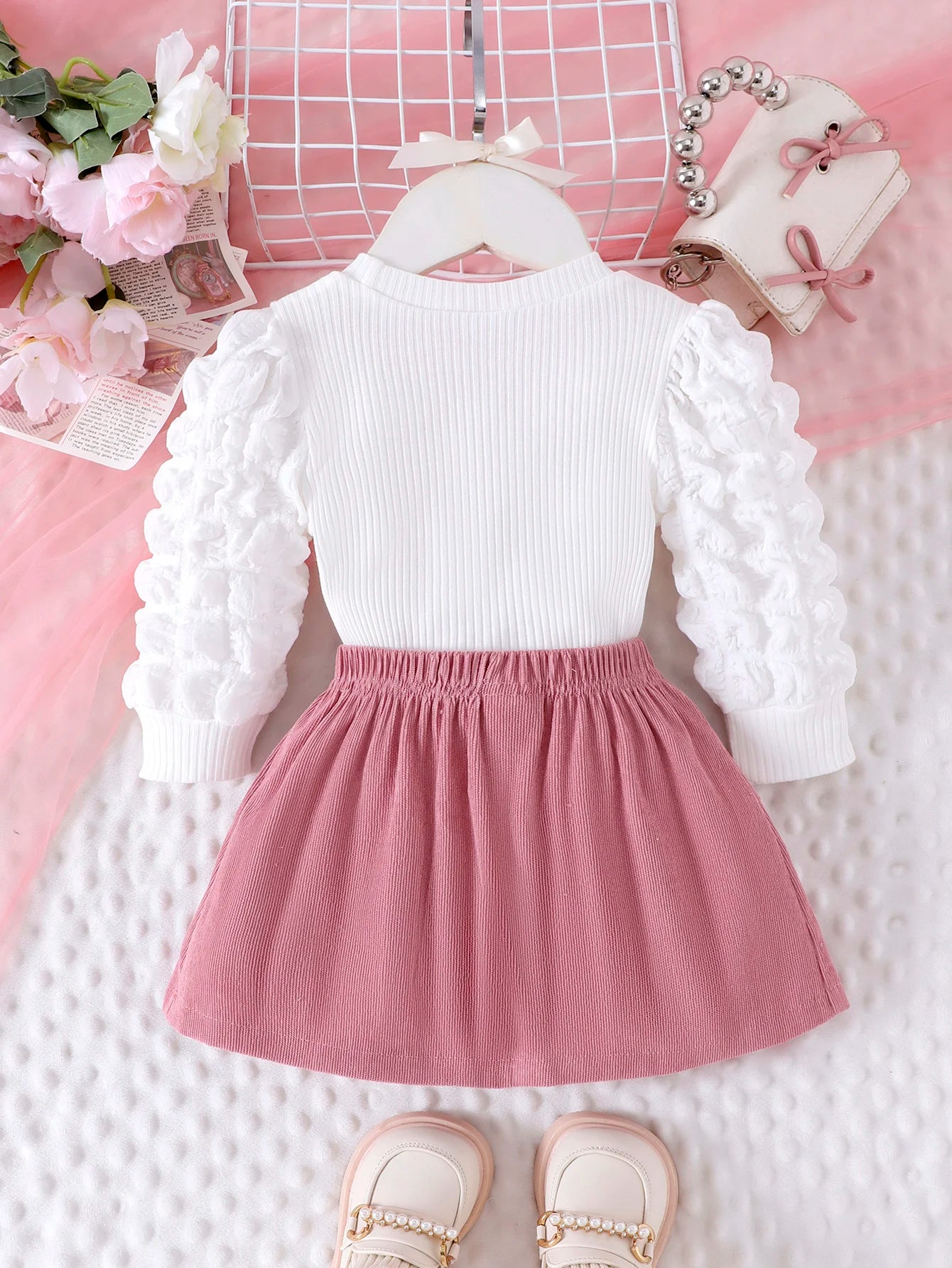 White long-sleeve top with ruffled sleeves and a pink skirt on a textured surface with decorative elements.